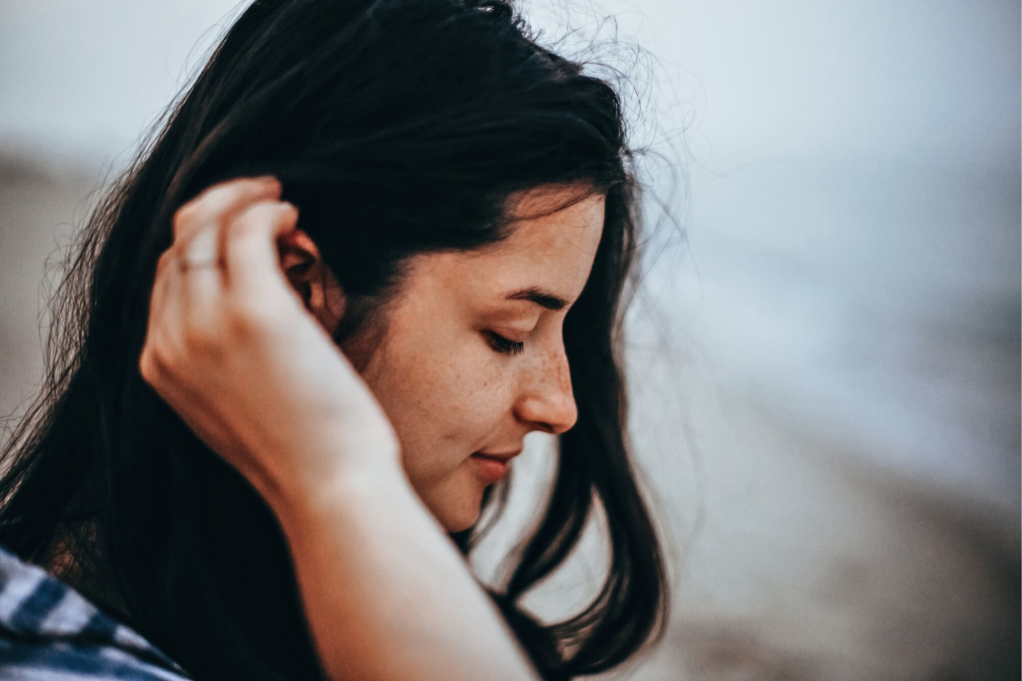 Close-up of a young woman with long dark hair, tilting her head slightly down while gently touching her ear with one hand. She has a serene expression and appears to be outdoors, with a blurred background suggesting a beach or seaside location.