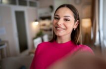 A young woman with long brown hair is taking a selfie indoors. She is smiling and wearing a bright pink sweater. The background shows a blurred interior of a cozy room, featuring soft lighting, a door, and some furnishings.