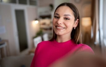 A young woman with long brown hair is taking a selfie indoors. She is smiling and wearing a bright pink sweater. The background shows a blurred interior of a cozy room, featuring soft lighting, a door, and some furnishings.