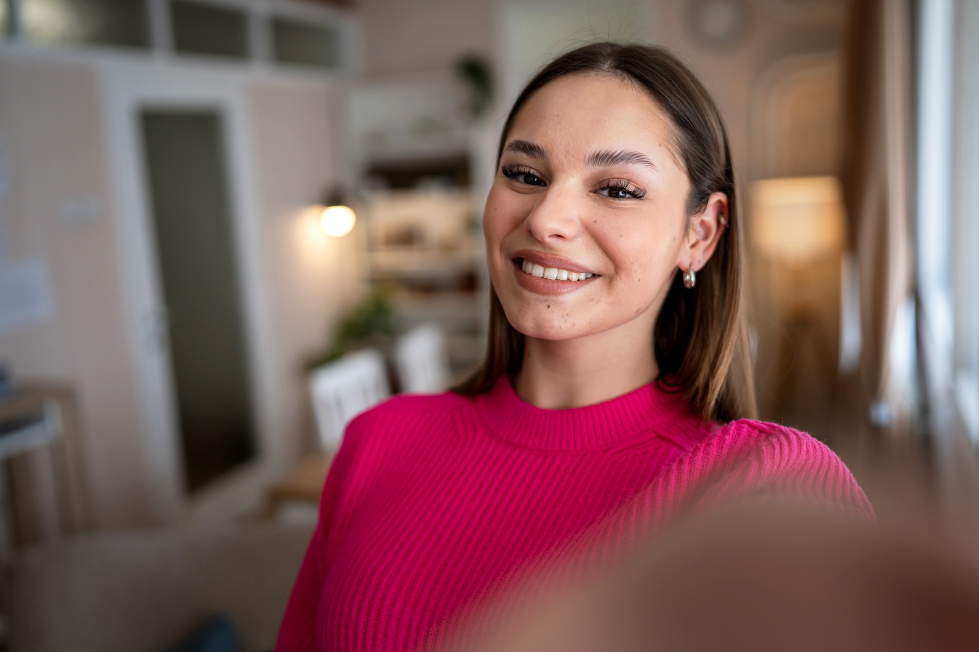 A young woman with long brown hair is taking a selfie indoors. She is smiling and wearing a bright pink sweater. The background shows a blurred interior of a cozy room, featuring soft lighting, a door, and some furnishings.