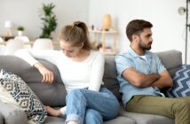 A young woman and man sit on a couch, facing away from each other with their arms crossed. The woman gazes downward, looking upset, while the man looks in another direction, also appearing displeased. The room has modern decor with plants and shelves in the background.