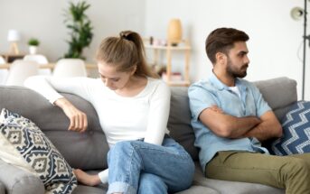 A young woman and man sit on a couch, facing away from each other with their arms crossed. The woman gazes downward, looking upset, while the man looks in another direction, also appearing displeased. The room has modern decor with plants and shelves in the background.