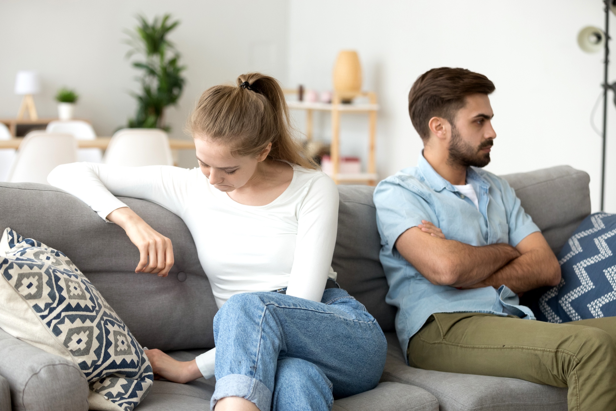 A young woman and man sit on a couch, facing away from each other with their arms crossed. The woman gazes downward, looking upset, while the man looks in another direction, also appearing displeased. The room has modern decor with plants and shelves in the background.