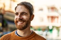 A bearded man wearing a brown sweater smiles while standing outdoors. The background is slightly blurred, showing buildings and a sunny day.