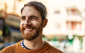 A bearded man wearing a brown sweater smiles while standing outdoors. The background is slightly blurred, showing buildings and a sunny day.