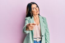 A woman with long dark hair wears a light green blazer over a beige top, paired with jeans. She appears to be pointing towards the camera with an expressive look on her face, standing against a pink background.