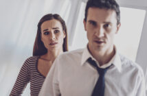 A woman with a concerned expression stands in the background, wearing a striped top. In the foreground, a man in a white dress shirt and loosened tie looks forward with a serious expression. Both are indoors with bright lighting.