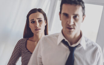 A woman with a concerned expression stands in the background, wearing a striped top. In the foreground, a man in a white dress shirt and loosened tie looks forward with a serious expression. Both are indoors with bright lighting.