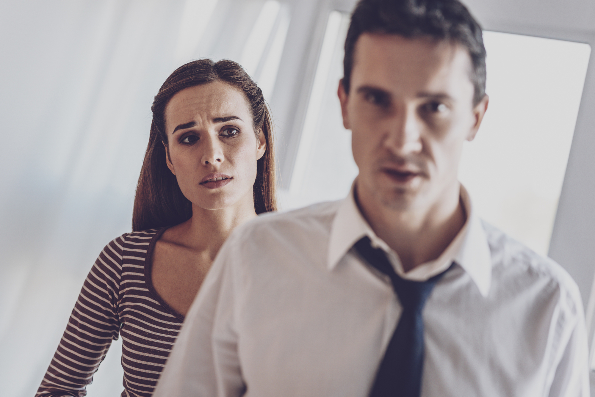 A woman with a concerned expression stands in the background, wearing a striped top. In the foreground, a man in a white dress shirt and loosened tie looks forward with a serious expression. Both are indoors with bright lighting.