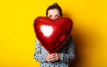 A person wearing a gray polka-dotted sweater is holding a red heart-shaped balloon in front of their face, peeking over the top. The background is a solid bright yellow, giving a cheerful and vibrant atmosphere.