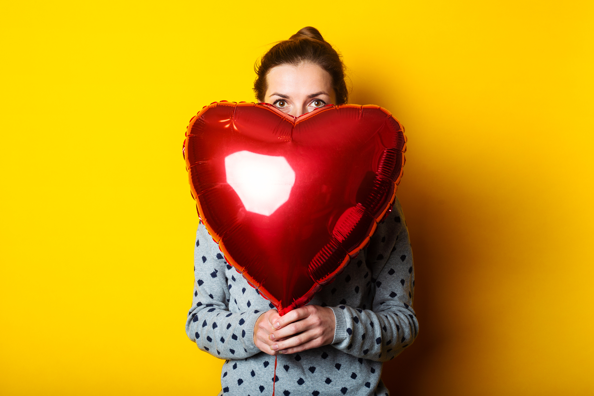 A person wearing a gray polka-dotted sweater is holding a red heart-shaped balloon in front of their face, peeking over the top. The background is a solid bright yellow, giving a cheerful and vibrant atmosphere.