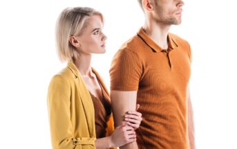 A woman with short blonde hair and wearing a yellow jacket is standing close to a man with short, light hair who is wearing a brown shirt. The woman is holding the man's arm, and both are looking to the right. The background is white and well-lit.