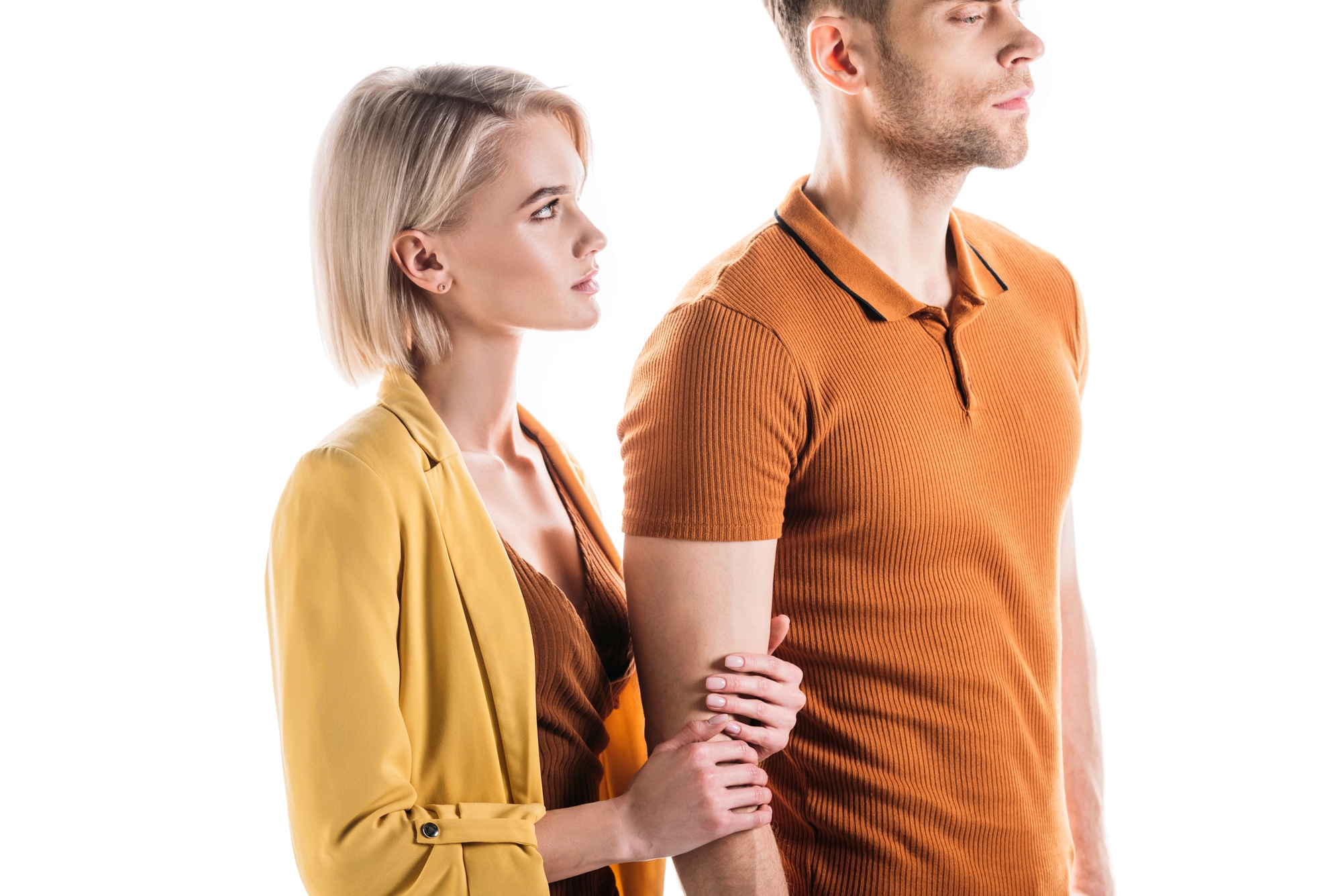 A woman with short blonde hair and wearing a yellow jacket is standing close to a man with short, light hair who is wearing a brown shirt. The woman is holding the man's arm, and both are looking to the right. The background is white and well-lit.