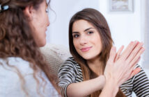 Two women are seated and smiling at each other. One woman, with long wavy hair, is facing away, while the other, with straight hair, is facing the camera, extending her hand forward as if giving a high-five. They are indoors and appear to be enjoying a conversation.