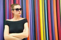 A woman with dark sunglasses and a black off-the-shoulder top stands with arms crossed in front of a colorful backdrop made of vertical, multicolored stripes. She has a serious expression and wears her hair in a bun.