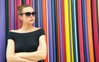 A woman with dark sunglasses and a black off-the-shoulder top stands with arms crossed in front of a colorful backdrop made of vertical, multicolored stripes. She has a serious expression and wears her hair in a bun.