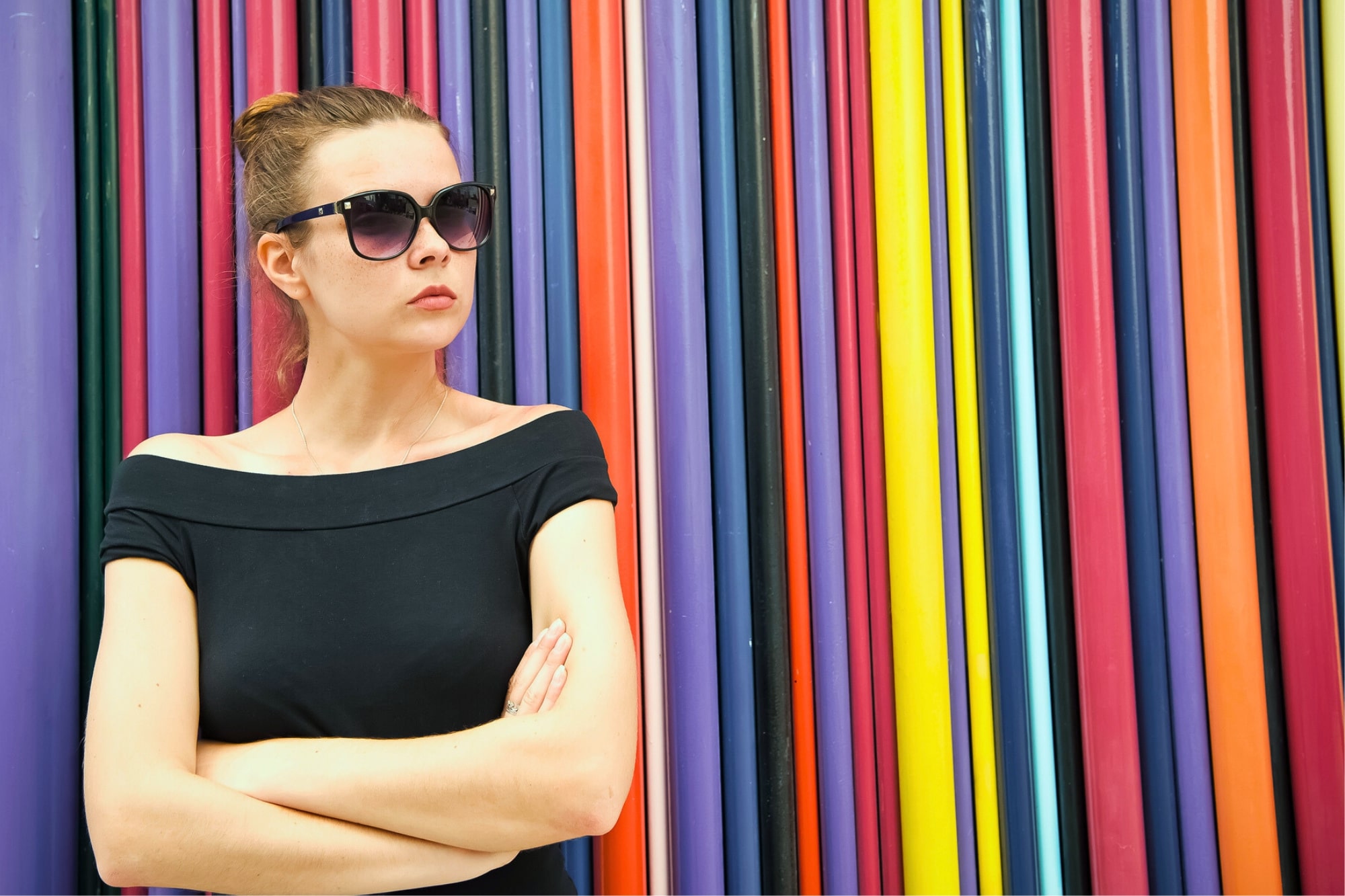 A woman with dark sunglasses and a black off-the-shoulder top stands with arms crossed in front of a colorful backdrop made of vertical, multicolored stripes. She has a serious expression and wears her hair in a bun.