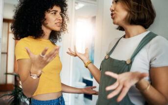 Two women are having an intense conversation in a brightly lit room. The woman on the left, with curly hair and wearing a yellow crop top, has a concerned expression. The woman on the right, with straight hair and wearing a white shirt under a green overall dress, gestures emphatically.