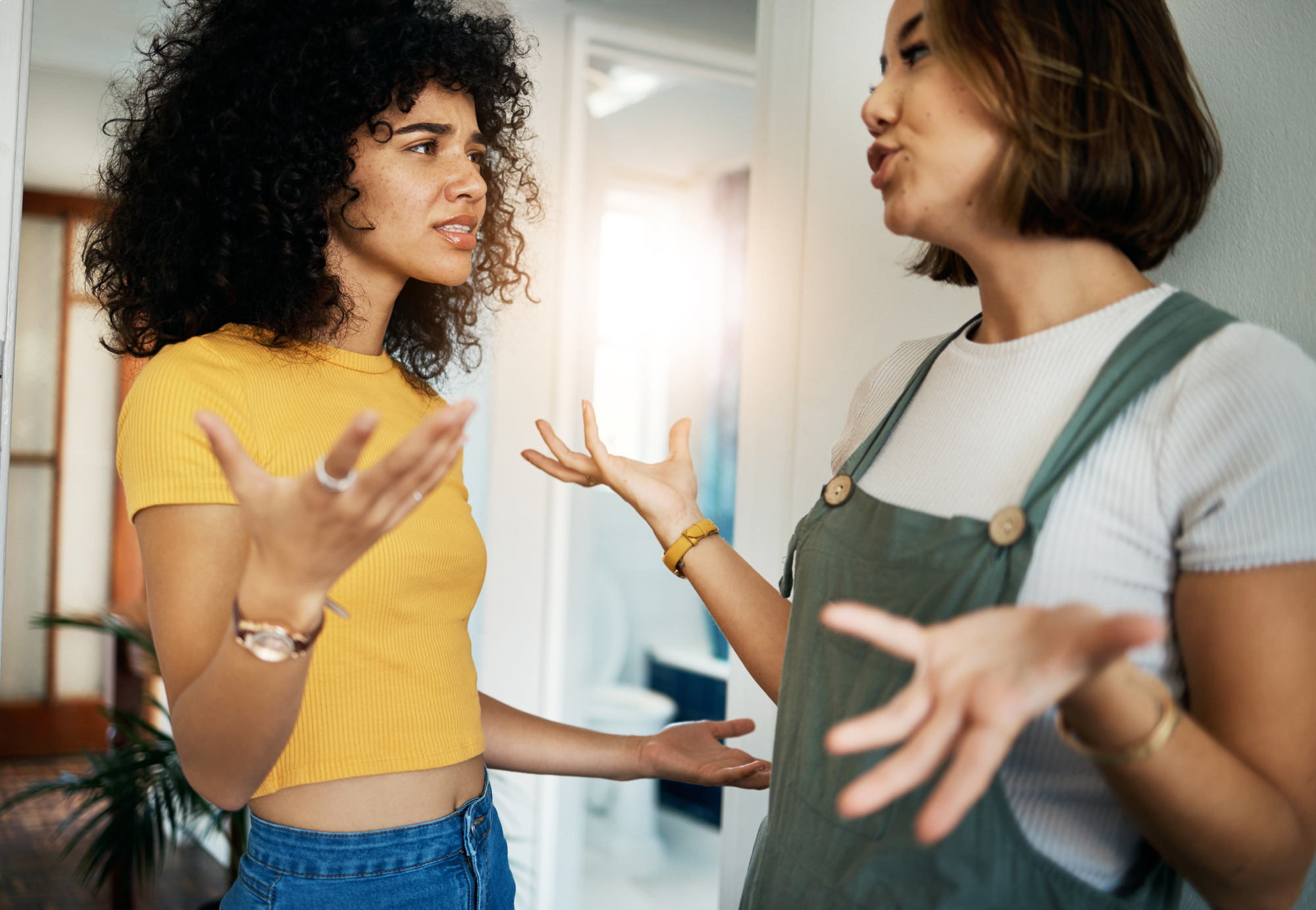 Two women are having an intense conversation in a brightly lit room. The woman on the left, with curly hair and wearing a yellow crop top, has a concerned expression. The woman on the right, with straight hair and wearing a white shirt under a green overall dress, gestures emphatically.