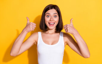 A woman with shoulder-length brown hair is smiling broadly and giving two thumbs up. She is wearing a white tank top and standing against a bright yellow background.