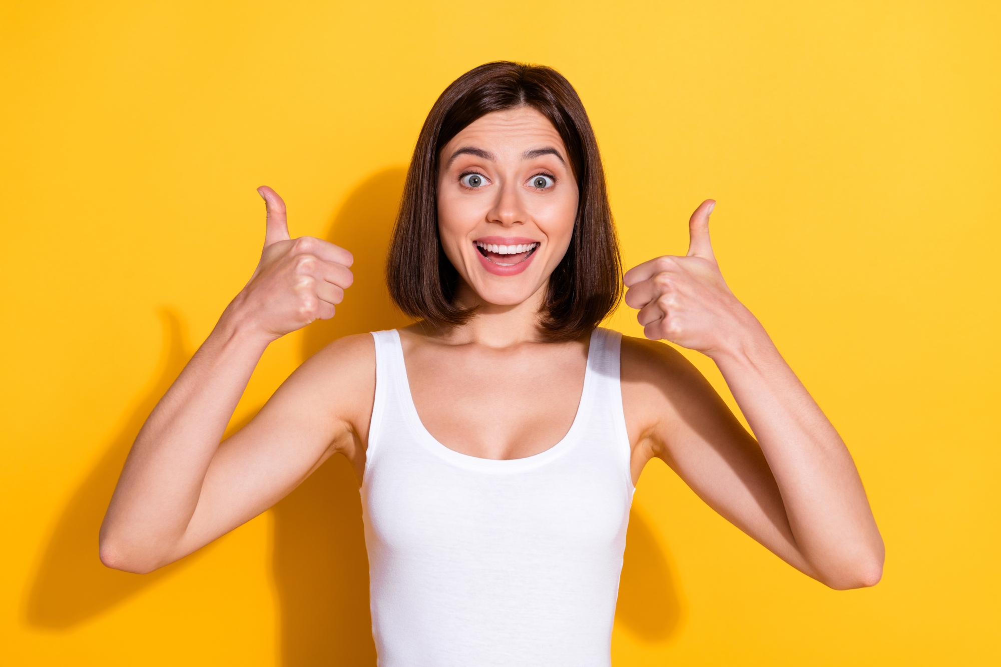 A woman with shoulder-length brown hair is smiling broadly and giving two thumbs up. She is wearing a white tank top and standing against a bright yellow background.