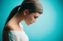 A woman with long brown hair tied back in a ponytail with a pink hair tie is shown in profile against a turquoise background. She is looking downwards and wearing an off-the-shoulder white top. The image has a soft and serene feel.