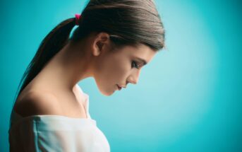 A woman with long brown hair tied back in a ponytail with a pink hair tie is shown in profile against a turquoise background. She is looking downwards and wearing an off-the-shoulder white top. The image has a soft and serene feel.