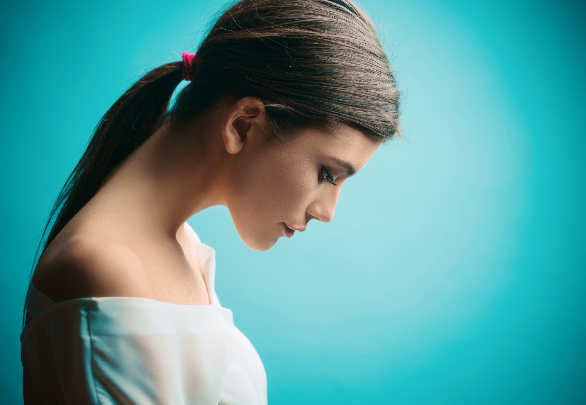 A woman with long brown hair tied back in a ponytail with a pink hair tie is shown in profile against a turquoise background. She is looking downwards and wearing an off-the-shoulder white top. The image has a soft and serene feel.