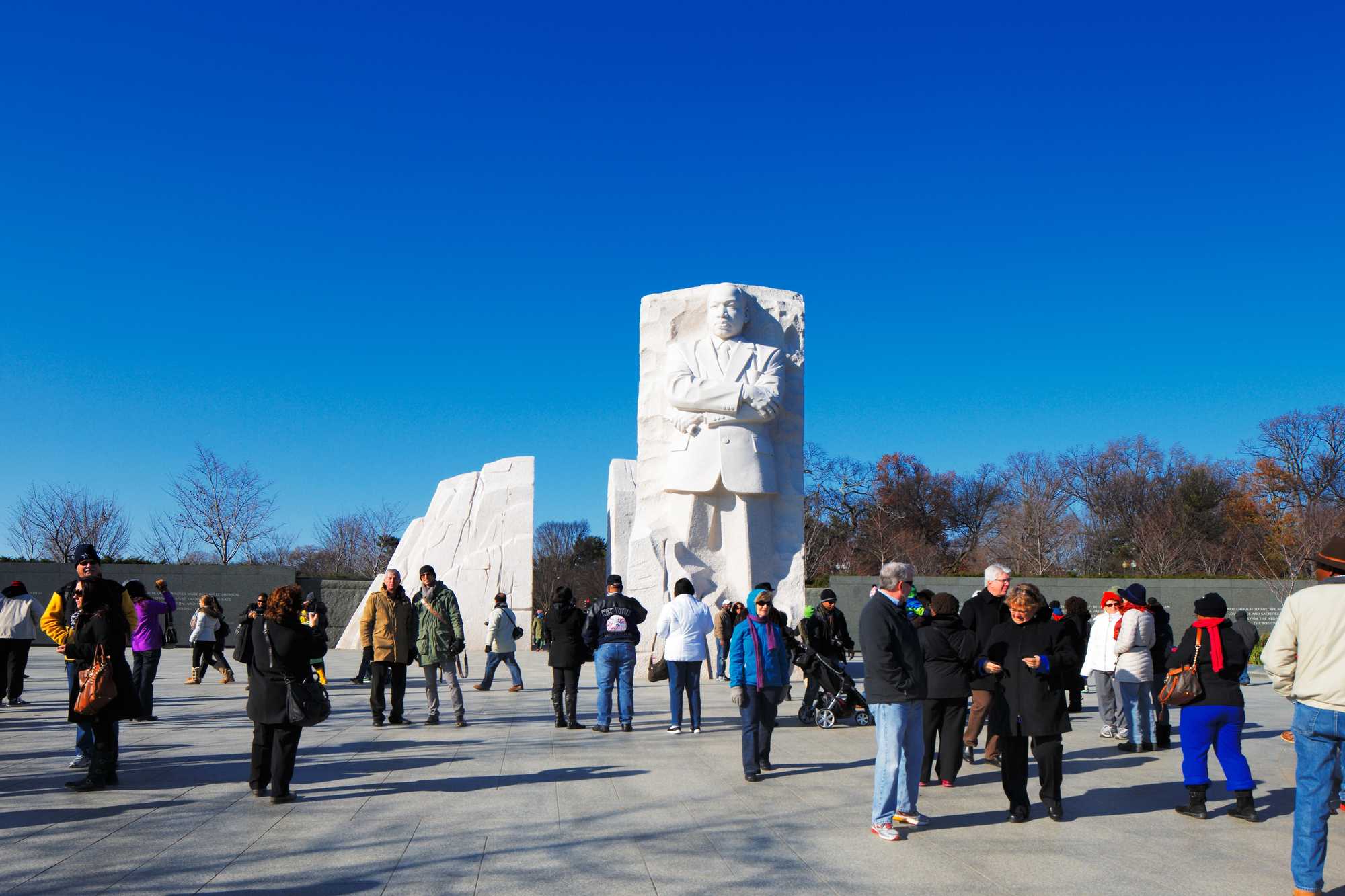 A diverse group of people stand and walk around the Martin Luther King Jr. Memorial on a clear day. The large stone statue of Martin Luther King Jr. with crossed arms is prominently displayed against a bright blue sky.