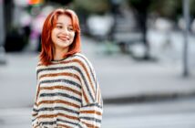 A woman with shoulder-length red hair and wearing a striped sweater is smiling as she stands on a city street. The background is blurred, showing vague outlines of buildings and street elements. The feel is casual and candid.