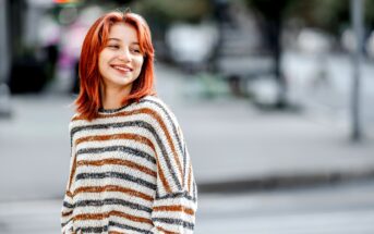 A woman with shoulder-length red hair and wearing a striped sweater is smiling as she stands on a city street. The background is blurred, showing vague outlines of buildings and street elements. The feel is casual and candid.