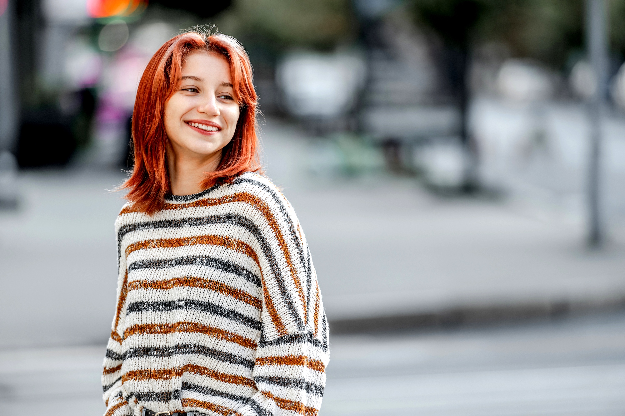 A woman with shoulder-length red hair and wearing a striped sweater is smiling as she stands on a city street. The background is blurred, showing vague outlines of buildings and street elements. The feel is casual and candid.