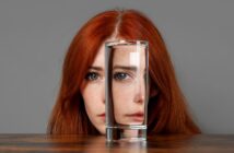 A woman with long red hair is partially obscured by a glass of water placed in front of her on a table. The water refracts and distorts her facial features, creating an intriguing visual effect. She is set against a neutral grey background.