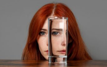 A woman with long red hair is partially obscured by a glass of water placed in front of her on a table. The water refracts and distorts her facial features, creating an intriguing visual effect. She is set against a neutral grey background.