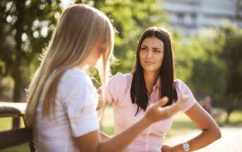 Two women are sitting on a bench in a park. One woman with light hair gestures as she speaks, while the other woman with dark hair listens with a serious expression. Trees and a building are visible in the background under sunny weather.