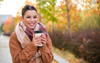 A woman with dark hair, wearing a brown jacket and a colorful scarf, smiles while holding a coffee cup. She stands on a leaf-covered path surrounded by vibrant autumn foliage. The background is slightly blurred, highlighting the fall colors.