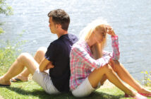 A man and a woman sit back-to-back on grass near a body of water. The man appears upset, staring into the distance, while the woman looks distressed, resting her head on her hand. They both wear casual clothes, including shorts and short-sleeve tops.