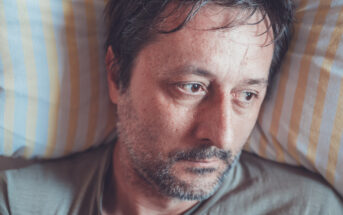 A close-up of a man with short dark hair and a beard lying on a yellow and white striped pillow, looking contemplative and somewhat sad. He is wearing a casual grey shirt, and his eyes seem to be focused on something outside the frame.