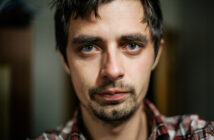 A close-up image of a man with short, dark hair and light eyes. He has a serious expression, light facial stubble, and wears a red and white plaid shirt. The background is blurred, focusing attention on his face.