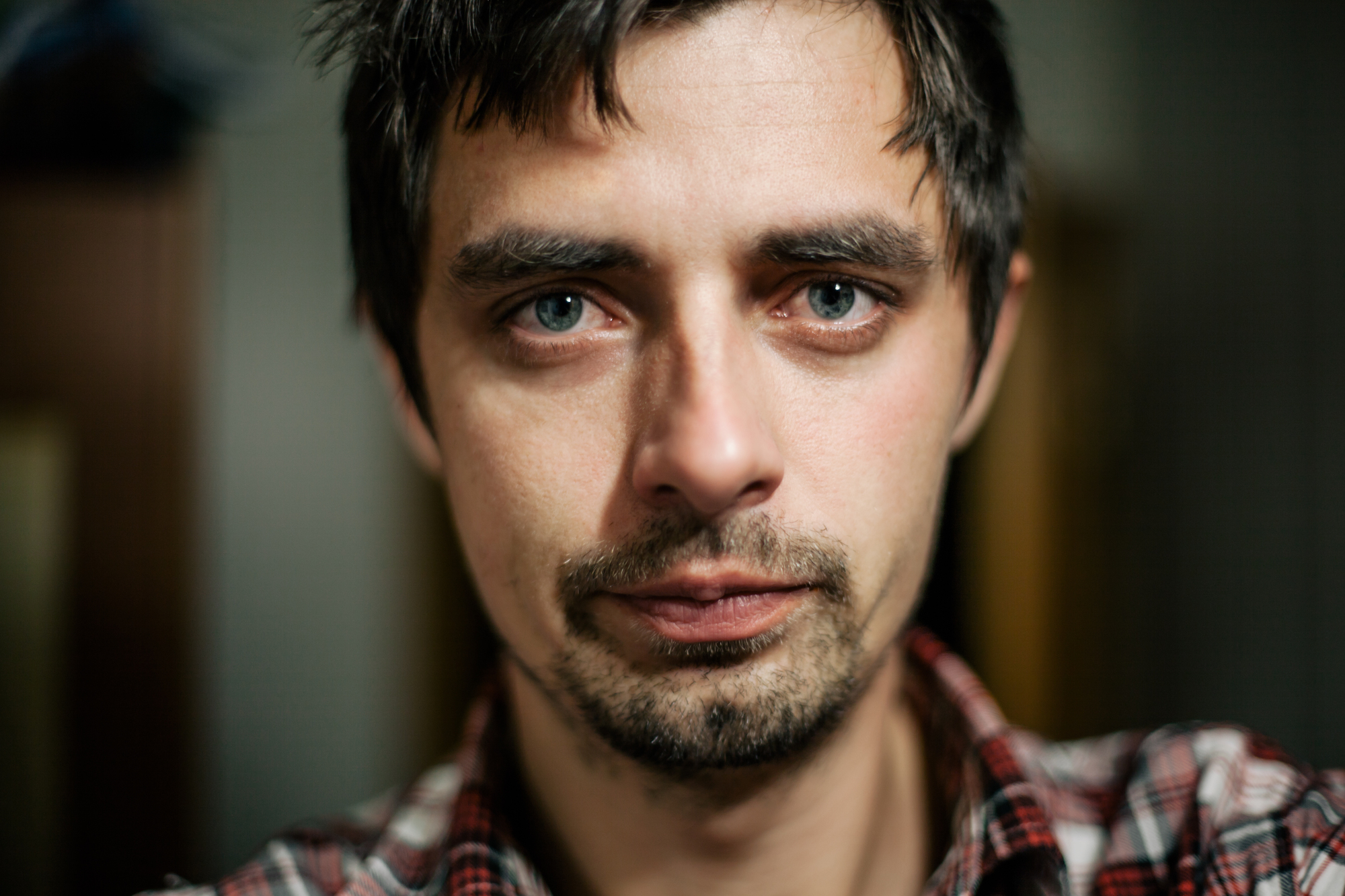 A close-up image of a man with short, dark hair and light eyes. He has a serious expression, light facial stubble, and wears a red and white plaid shirt. The background is blurred, focusing attention on his face.