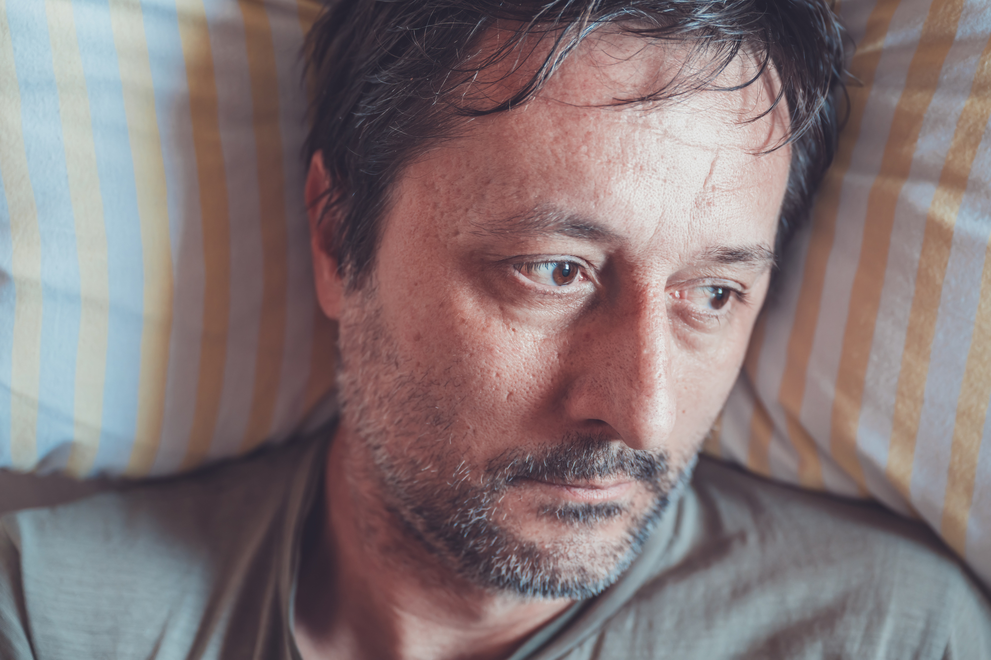 A close-up of a man with short dark hair and a beard lying on a yellow and white striped pillow, looking contemplative and somewhat sad. He is wearing a casual grey shirt, and his eyes seem to be focused on something outside the frame.