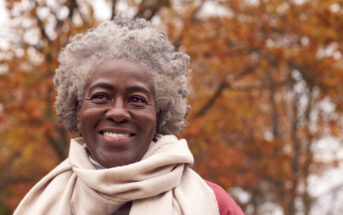 A smiling person with short curly gray hair is wearing a light-colored scarf and jacket. The background features colorful autumn trees with red, orange, and yellow leaves, creating a warm and vibrant setting.