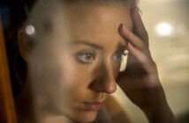 A woman with a distressed expression rests her forehead on her hand. She is seen through a glass, which partially blurs the image. The soft lighting and close-up focus highlight her contemplative demeanor.