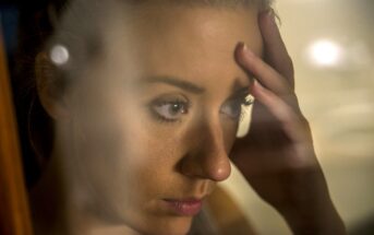 A woman with a distressed expression rests her forehead on her hand. She is seen through a glass, which partially blurs the image. The soft lighting and close-up focus highlight her contemplative demeanor.