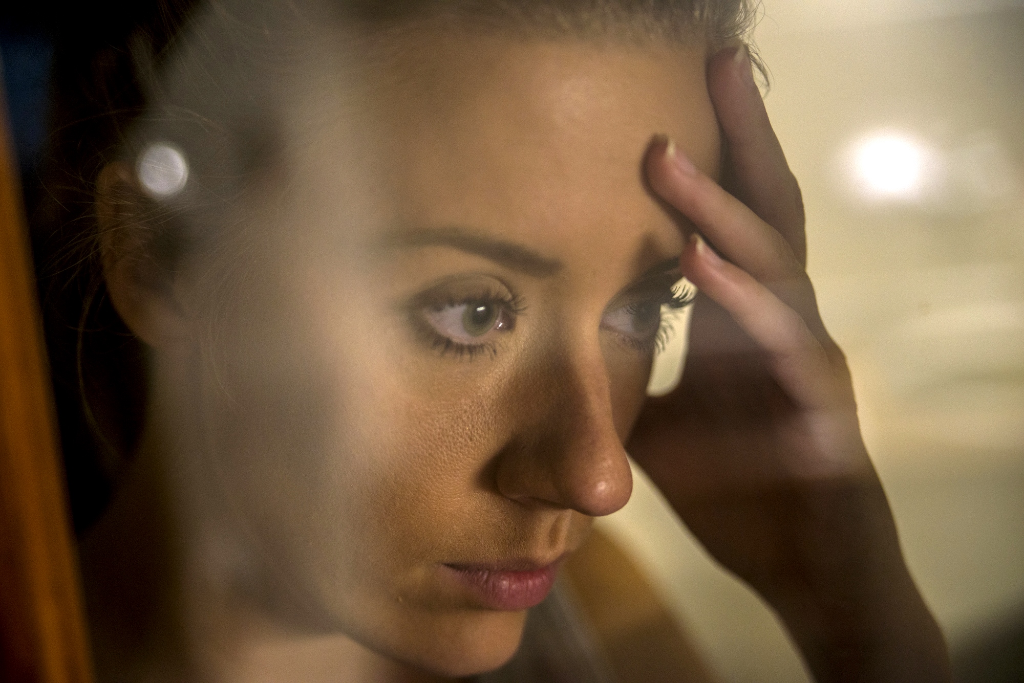 A woman with a distressed expression rests her forehead on her hand. She is seen through a glass, which partially blurs the image. The soft lighting and close-up focus highlight her contemplative demeanor.