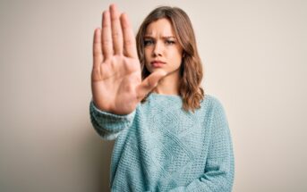 A young woman with shoulder-length brown hair wearing a light blue sweater is standing against a beige background. She has a serious expression on her face and is holding her hand up in a stopping gesture directly in front of her.