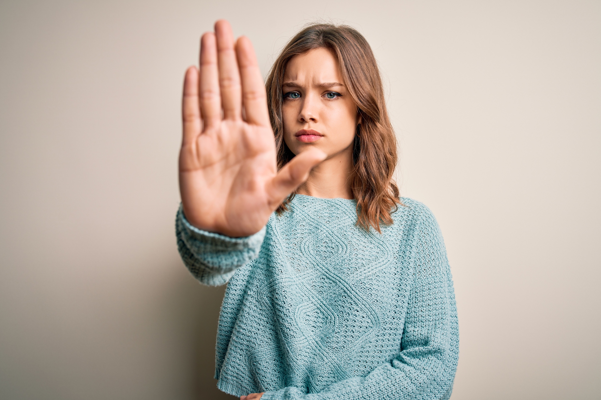 A young woman with shoulder-length brown hair wearing a light blue sweater is standing against a beige background. She has a serious expression on her face and is holding her hand up in a stopping gesture directly in front of her.