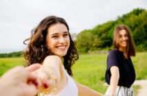 Two young women smile as they walk hand-in-hand through a lush, green field. The woman in the foreground, with curly hair and a white tank top, faces the camera, while the woman behind her with straight hair and a black shirt looks to the side. Trees are in the background.