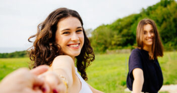 Two young women smile as they walk hand-in-hand through a lush, green field. The woman in the foreground, with curly hair and a white tank top, faces the camera, while the woman behind her with straight hair and a black shirt looks to the side. Trees are in the background.