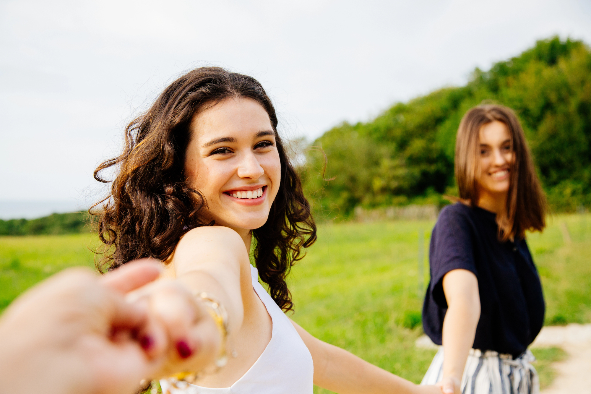 Two young women smile as they walk hand-in-hand through a lush, green field. The woman in the foreground, with curly hair and a white tank top, faces the camera, while the woman behind her with straight hair and a black shirt looks to the side. Trees are in the background.